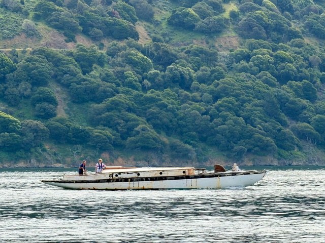 Yankee's bare hull under tow across San Francisco Bay, Marin hills behind